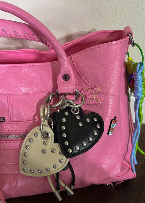 A close-up of a pink textured leather handbag with two heart-shaped keychains attached. One keychain is cream-colored with silver studs, and the other is black with silver studs.