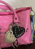 A close-up of a pink textured leather handbag with two heart-shaped keychains attached. One keychain is cream-colored with silver studs, and the other is black with silver studs.