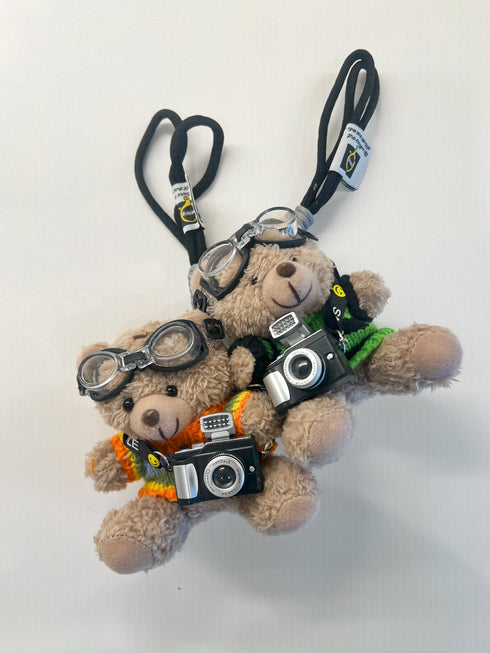 Two plush teddy bear keychains styled as photographers, wearing round aviator-style goggles, black neck lanyards, and holding miniature black-and-silver cameras. One bear wears an orange and yellow striped shirt, and the other wears a green shirt.