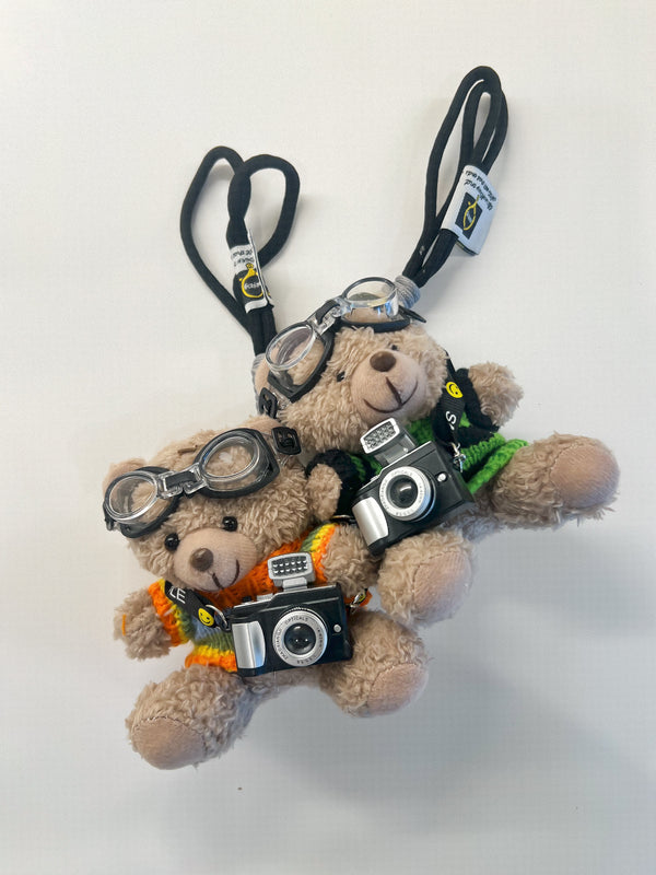 Two plush teddy bear keychains styled as photographers, wearing round aviator-style goggles, black neck lanyards, and holding miniature black-and-silver cameras. One bear wears an orange and yellow striped shirt, and the other wears a green shirt.