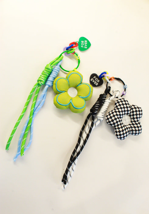 Two handmade braided rope keychains with flower-shaped charms on a plain white background. One keychain features green and light blue braided ropes with a lime green flower charm.