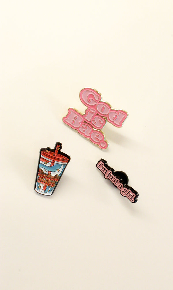 Three enamel pins displayed on a light cream background: a pink 'God Is Bae.' pin, a colorful coffee cup pin, and a black pin with pink text reading 'im just a girl!'.
