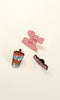 Three enamel pins displayed on a light cream background: a pink 'God Is Bae.' pin, a colorful coffee cup pin, and a black pin with pink text reading 'im just a girl!'.