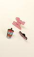 Three enamel pins displayed on a light cream background: a pink 'God Is Bae.' pin, a colorful coffee cup pin, and a black pin with pink text reading 'im just a girl!'.